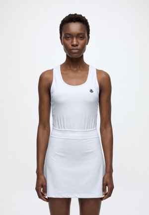 Femme debout, face à l’avant, portant une robe de sport blanche sans manches avec un petit logo sur la poitrine et un détail en mesh à la taille.