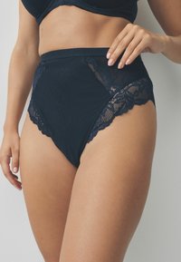 Hochtaillierte schwarze Panties aus glattem Stoff mit Spitze, die gewellte Kanten und durchsichtige Einsätze an den Seiten aufweisen.