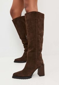 Stivali in suede marrone alti fino al ginocchio con punta quadrata, tacco a blocco spesso e suola in gomma, caratterizzati da un dettaglio di zip laterale.