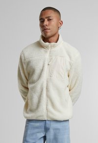 Giacca in pile bianca con colletto alto e chiusura con zip, dotata di tasca sul petto beige, superficie testurizzata e vestibilità rilassata.