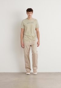 Beige t-skjorte med korte ermer og lys tekst, kombinert med beige bukser og lyse sneakers. Stoffet ser jevnt og uformelt ut.