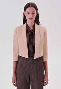 Bijgesneden beige blazer met een gladde textuur, brede revers en driekwart mouwen, gecombineerd met een bruine blouse en bruine broek met patroon.