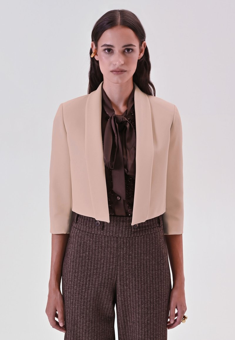 Bijgesneden beige blazer met een gladde textuur, brede revers en driekwart mouwen, gecombineerd met een bruine blouse en bruine broek met patroon.