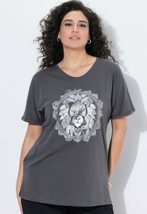 Frau mit lockigem Haar, die ein dunkelgraues T-Shirt mit detailliertem weißem Löwengesicht und dekorativem kreisförmigen Muster trägt, vor weißem Hintergrund stehend.