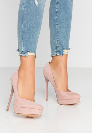 Ben i lyseblå revnede skinny jeans iført blegrøde ruskind platform høje hæle på en hvid gulv.