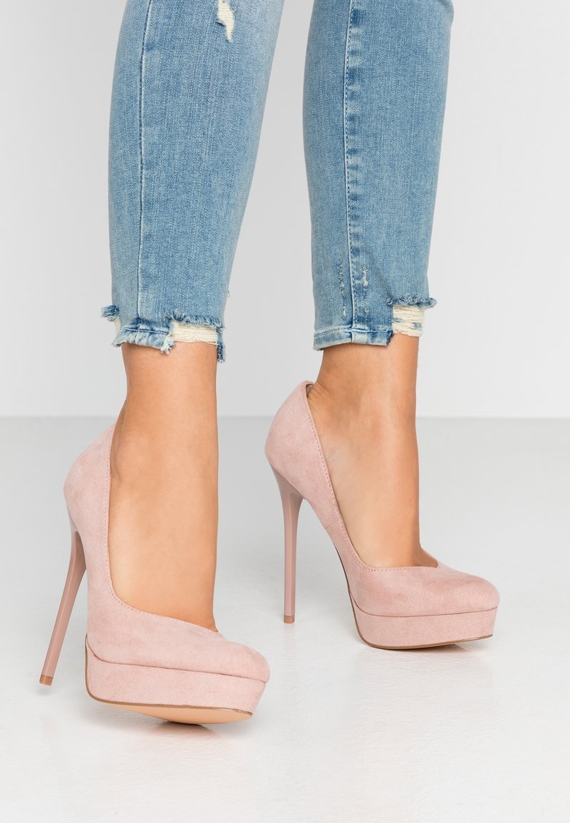 Jambes en jean skinny bleu clair déchiré portant des talons compensés en daim rose pâle sur un sol blanc.