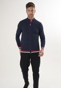 Dunkelblauer Reißverschluss-Strickcardigan mit einem strukturierten Muster, ausgestattet mit rot-weißen gestreiften Akzenten am Kragen und Saum, kombiniert mit schwarzen Hosen.