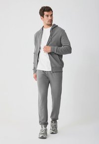 Grijze hoodie met rits en bijpassende joggingbroek van zachte, gestructureerde stof, gedragen over een wit T-shirt. Grijze sportieve schoenen maken de outfit compleet.