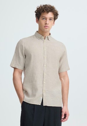 Mlad moški z kodrastimi lasmi, oblečen v bež srajco s kratkimi rokavi in črne hlače, stoji z eno roko v žepu.