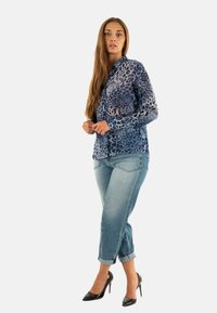 Vrouw die staat in een blauwe luipaardprint blouse, opgerolde lichtblauwe jeans en zwarte hoge hakken op een witte achtergrond.