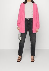 Pink fuzzy oversized cardigan with dropped shoulders, paired with a ribbed light grey top, black high-waisted jeans, and strappy block-heel sandals.