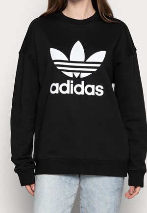 Person iført en sort sweatshirt med et stort hvidt Adidas trefoil-logo og tekst på fronten, kombineret med lyseblå jeans.