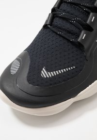 Primo piano di una sneaker nera Nike con punta testurizzata, il logo swoosh Nike a strisce e lacci neri con macchie bianche, su sfondo bianco.