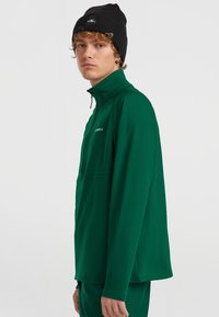 Grüne, nahtlose, langärmlige Jacke mit Stehkragen; aus glattem, dehnbarem Stoff. Schwarze Strickmütze mit einem Logotag.