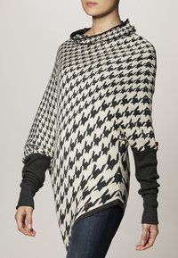 Zwart-wit houndstooth patroon poncho met een colhals en lange zwarte mouwen. Heeft een bruin merkknopdetail.