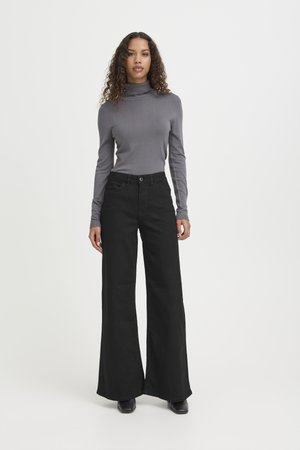 Femeie cu păr creț, purtând un pulover gri cu guler înalt, pantaloni negri largi cu talie ridicată și pantofi negri, stând pe un fundal alb.