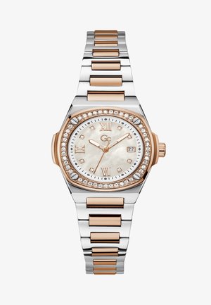Ezüst és rose gold színű óra kerek tokban, gyöngyház számlappal, gyémánt díszítésekkel és lánckarkötővel. Római számokkal és dátumkijelzővel rendelkezik.