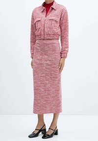 Femme portant une veste à carreaux rose et blanc assortie à une longue jupe, un haut rouge en dessous, et des chaussures Mary Jane noires à talons bas.