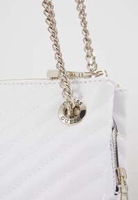 Sac à main en cuir blanc matelassé avec une bandoulière en chaîne dorée et une garniture circulaire dorée gravée de "GUESS."