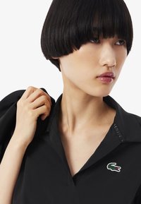Camisa polo negra con cuello en V y un sutil logo de cocodrilo. Textura suave, mangas cortas y una etiqueta bordada en el cuello.