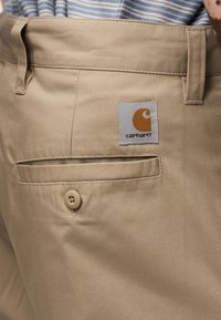 Μπεζ παντελόνι από λείο ύφασμα, με κλείσιμο με κουμπί, πίσω τσέπη και τετράγωνο μπαλώμα λογότυπο Carhartt.