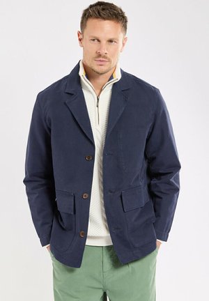 Homme portant un blazer bleu marine sur un pull blanc à zip court et un pantalon vert, debout les mains dans les poches devant un fond uni.