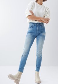 Jeans de tiro alto en azul con un acabado desgastado, de corte ajustado y con pasadores para cinturón. Combinados con botas de tobillo color beige claro y un sudadera color crema.