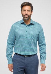 Een knoopshirt met een geometrisch patroon in turquoise, blauw en paars, met lange mouwen en een cutaway kraag. Draagt een blauwe broek.