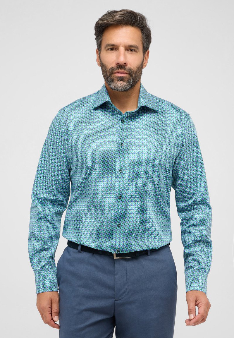 Een knoopshirt met een geometrisch patroon in turquoise, blauw en paars, met lange mouwen en een cutaway kraag. Draagt een blauwe broek.