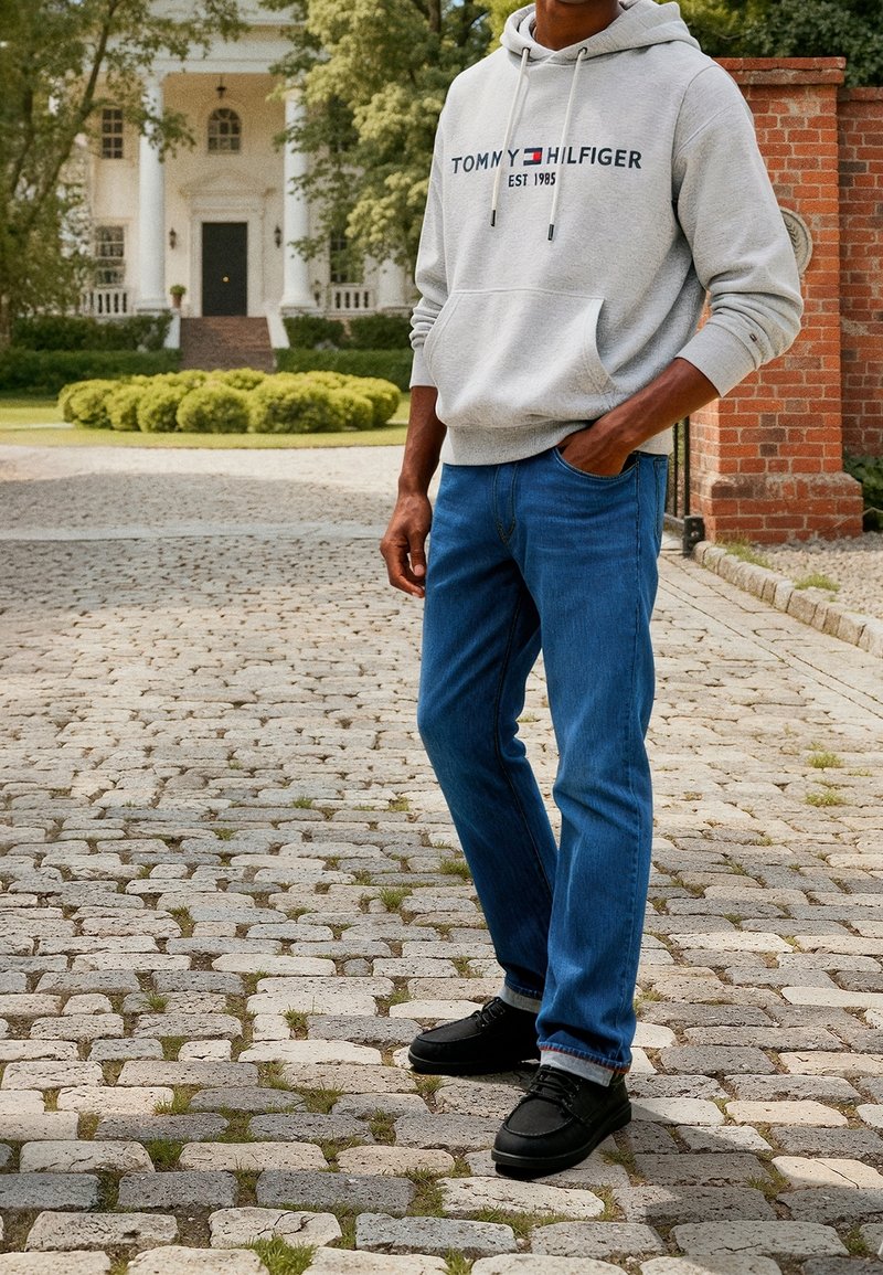 Muž v šedé mikině Tommy Hilfiger, modrých džínách a černých botách stojí na dlážděné cestě poblíž velké bílé budovy a zeleně.