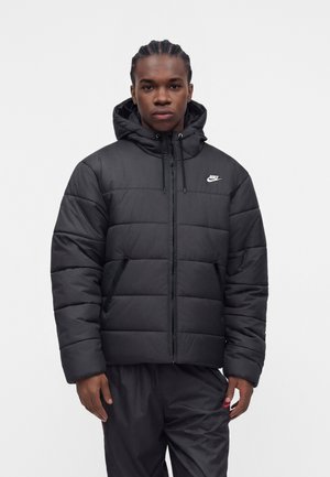 Junger Mann mit geflochtenen Haaren, der eine schwarze Nike gesteppte Jacke mit Kapuze und schwarze Hose trägt, steht vor einem schlichten hellen Hintergrund.