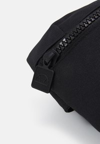 Musta kangaslaukku, jossa on teksturoitu vetoketjusuljenta ja neliönmuotoinen vedinlogo. Pinnan viimeistely on sileä ja mattapintainen.