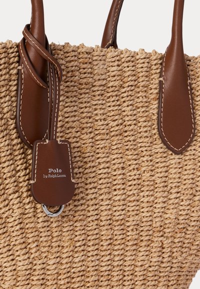 Fonott szalmakosár barna bőr fogantyúkkal és egy bőr címkével, amelyen a "Polo by Ralph Lauren" felirat olvasható, és az egyik fogantyú közelében van rögzítve.