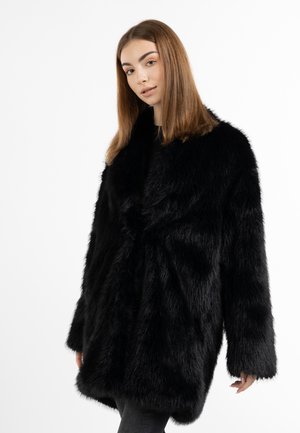 Jonge vrouw met steil bruin haar draagt een grote zwarte harige jas tegen een effen witte achtergrond.