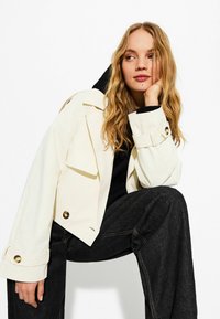Veste couleur crème avec une coupe décontractée, dotée de boutons dorés, de grandes poches avant et de manches larges. Porte sur un haut noir et un pantalon en denim.