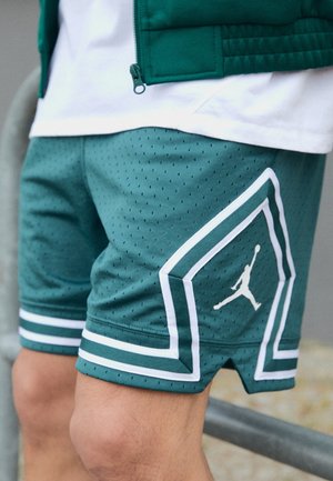 Shorts de sport bleu sarcelle avec bordure blanche et logo Jumpman, portés avec une veste à zip verte et un t-shirt blanc, montrant les jambes d'une personne en position médiane.