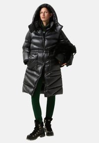 Černý nafukovací kabát s kapucí, zdobený prošívaným vzorem a předními knoflíky. Kombinován se zelenými sametovými kalhotami a černými botami s kožešinovou podšívkou.