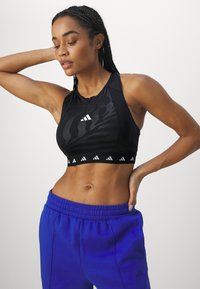 Svart atletisk crop topp med et teksturert mønster, hvit logo og uttalt bånd. Kombinert med blå joggebukser med elastisk midje.