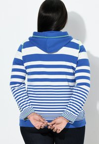 Osoba s dlouhými tmavými vlasy, oblečená v modrobílé pruhované mikině s kapucí, stojící se sepjatýma rukama za zády na bílém pozadí.