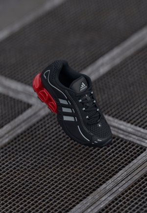 Basket Adidas noire avec des bandes grises et une semelle rouge amortissante, flottant au-dessus d'une surface métallique en grille.