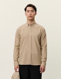 Šviesiai rudas ilgas rankovėmis mygtukų marškinis su apykakle, pasižymintis subtilia tekstūra ir priekiu kišene, derinamas su juodomis kelnėmis.