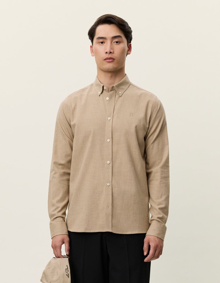 Šviesiai rudas ilgas rankovėmis mygtukų marškinis su apykakle, pasižymintis subtilia tekstūra ir priekiu kišene, derinamas su juodomis kelnėmis.