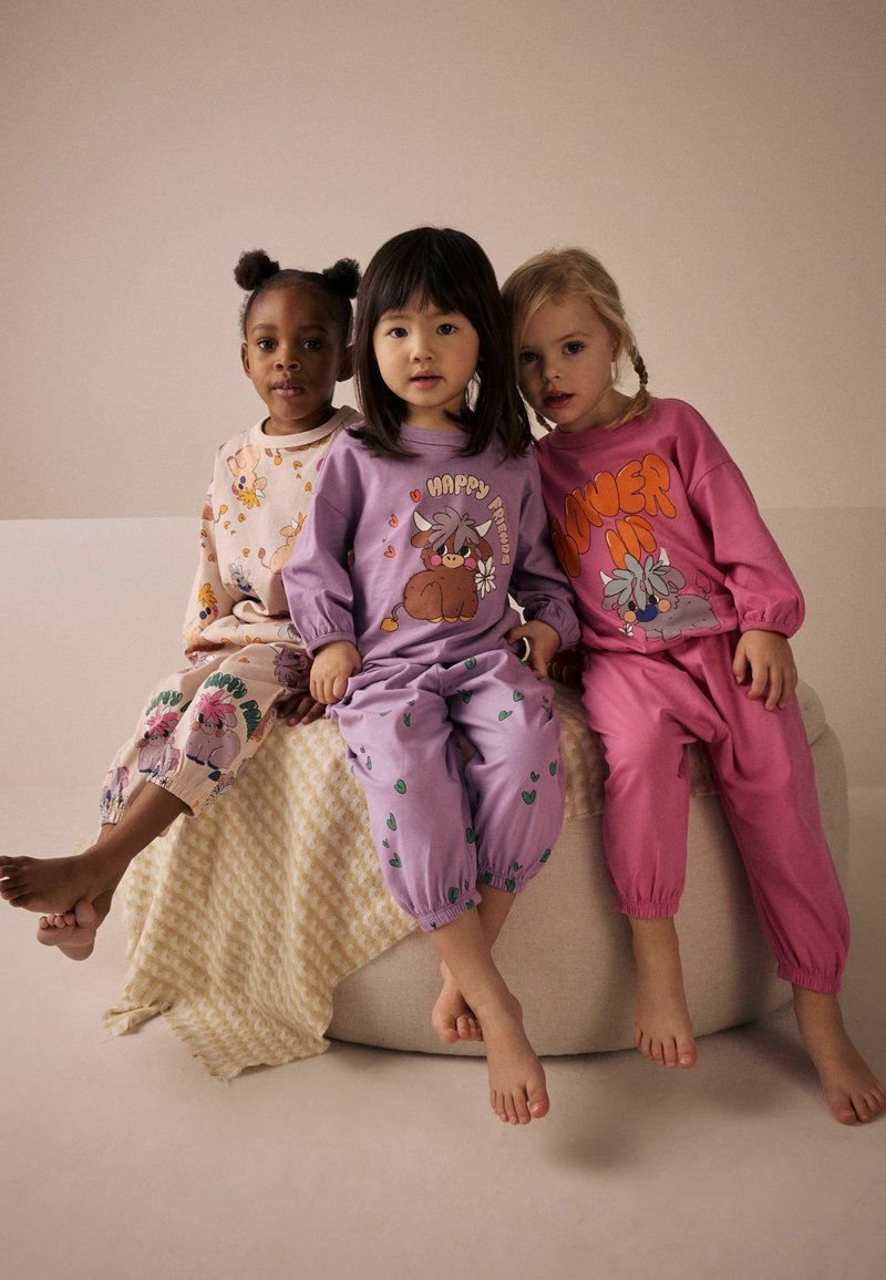 Drie kinderen zitten op een lichtgekleurde ronde kussen. Ze dragen kleurrijke loungewear: een crème top met dierenprints, een paarse outfit met een graphic, en een roze top.
