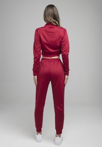 Rød træningsdragt med en cropped langærmet trøje og tapered bukser. Glat stof, elastisk talje og tætsiddende manchetter. Hvide sneakers.