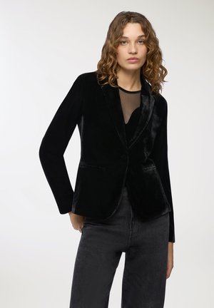 Blazer di velluto nero con rever in satin, chiusura a bottone singolo e due tasche frontali. Indossato sopra una maglia nera trasparente e abbinato a jeans scuri.