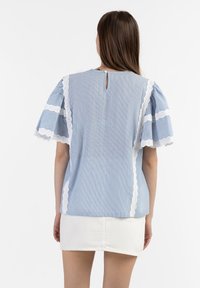 Una modella che indossa una blusa blu a righe con finiture in pizzo bianco e maniche a sbuffo, abbinata a una gonna di denim bianca, vista di spalle.