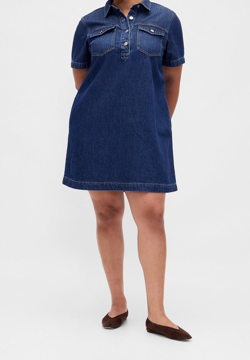 Personne portant une robe en denim bleu foncé à manches courtes avec col et boutons, associée à des chaussures plates marron foncé, debout sur un fond uni.