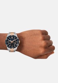 Kronograaf kell, millel on must siht, hõbedane roostevabast terasest ja roosakuldse aktsendiga, suure ümmarguse korpuse ja tekstuuriga rihm.