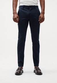 Chinos bleu marine en coton avec une coupe slim, dotés de poches standard et sans motifs visibles, associés à des mocassins marron.