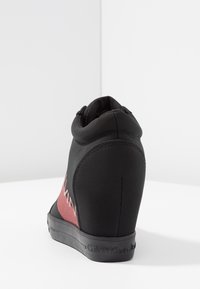 Svart kilsko med röd sidopanel och "Calvin Klein Jeans"-logotyp på sulan och sidan, fotograferad mot en vit bakgrund.